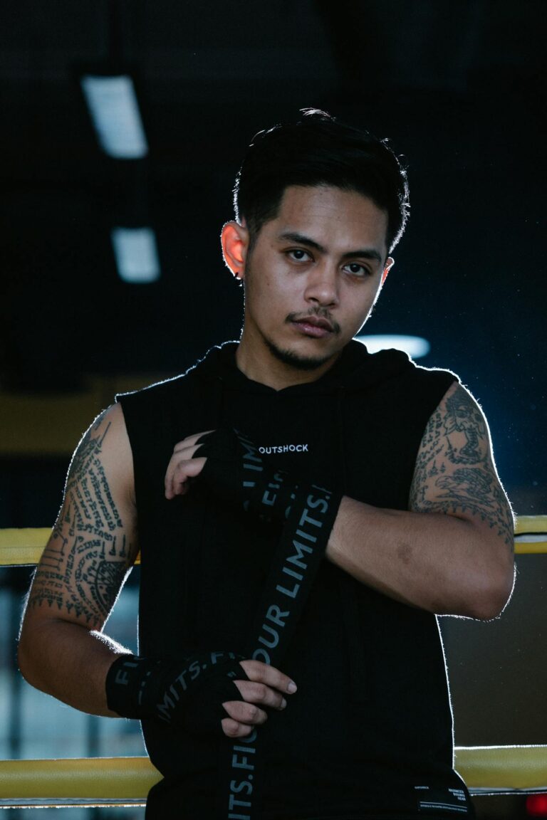 Serious ethnic male boxer in black t shirt looking at camera and wrapping bandage on hand against ring ropes